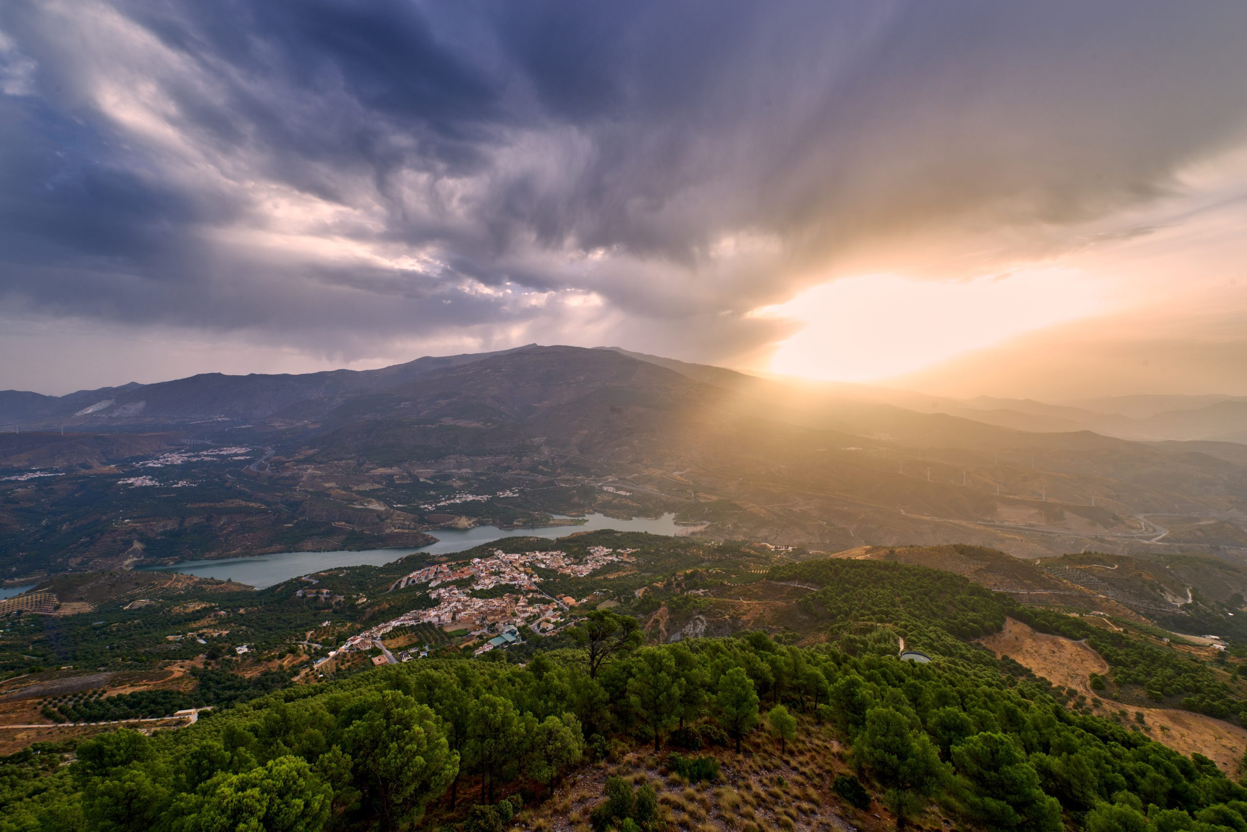 Vista aérea del Valle de Lecrín, en Granada, España, al atardecer. En la imagen se aprecia un paisaje montañoso con extensos pinares en primer plano, un pueblo blanco junto a un embalse en el centro, y montañas al fondo. 